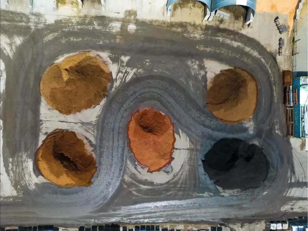 Aerial view of a landscaping supply yard with large piles of mulch and soil in different colors, including black, red, and natural brown, arranged on a concrete lot with equipment and storage buildings nearby.